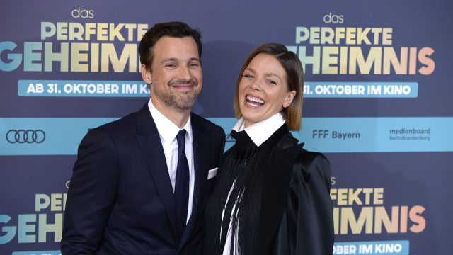 Auf diesem Bild zur symbolischen Ankündigung von Der perfekte Urlaub sind Florian David Fitz und Jessica Schwarz bei der Premiere von Das perfekte Geheimnis zu sehen. Beide Schauspieler lächeln herzlich in die Kamera. Fitz trägt einen dunklen Anzug mit Krawatte, während Schwarz mit weißem Hemd und schwarzem Blazer auffällt. Die Stimmung wirkt gelöst und fröhlich.