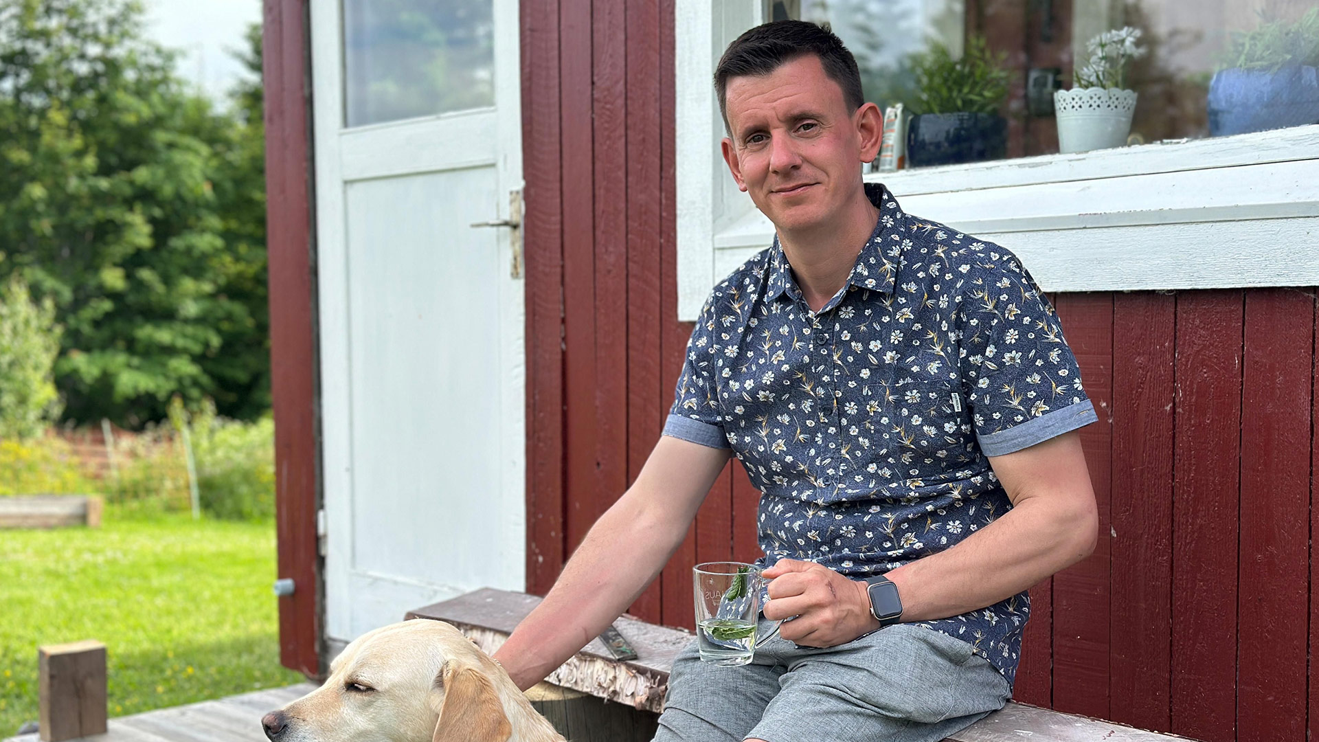 Auf dem Bild zu Bauer sucht Frau International 2026 sitzt ein Mann entspannt vor einem roten Holzhaus, hält ein Glas in der Hand und streichelt einen Hund.
