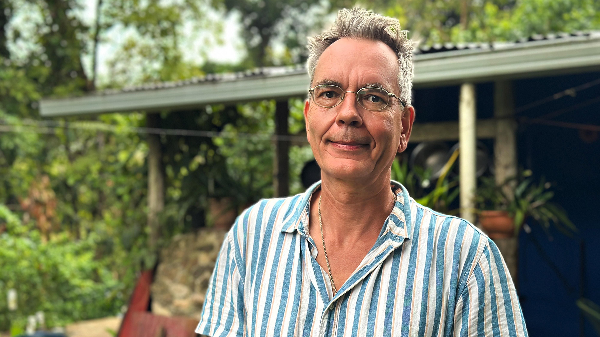 Auf dem Bild zu Bauer sucht Frau International 2026 steht ein Mann mit gestreiftem Hemd und Brille vor einem Haus mit Garten.