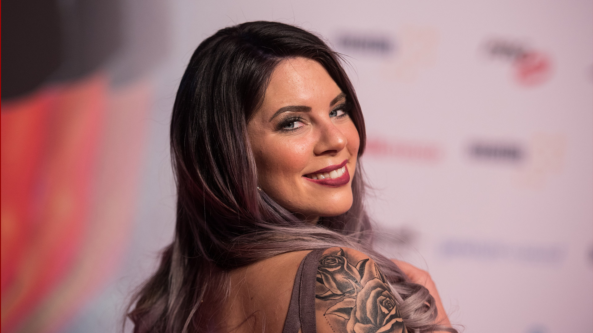 Auf dem Bild ist die Dschungelcamp-Gewinnerin Jenny Frankhauser zu sehen. Sie blickt lächelnd über die Schulter in die Kamera und trägt ein schulterfreies Outfit, das ein großes Rosen-Tattoo auf ihrem Oberarm zeigt. Ihre langen, gewellten Haare verlaufen in einem dunklen bis lilafarbenen Ton. Der Hintergrund zeigt eine Veranstaltung mit Logos und Sponsorenwänden.