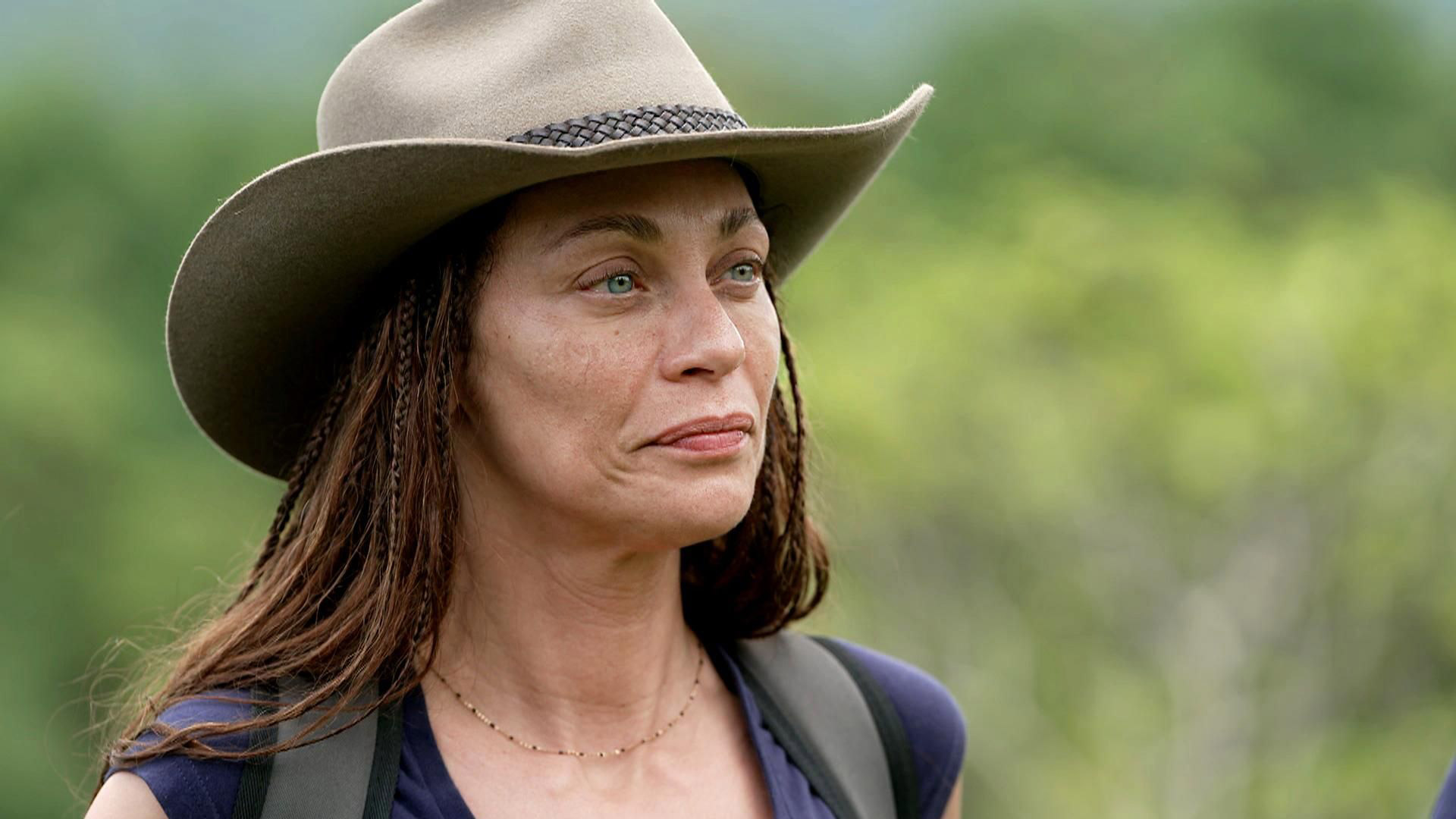 Auf dem Bild ist Dschungelcamp-Gewinnerin Lilly Becker in einem natürlichen Dschungel-Setting zu sehen. Sie trägt einen breitkrempigen Hut und schaut ernst, aber gefasst in die Ferne. Ihr Look ist natürlich, mit geflochtenen Haarsträhnen und einem Rucksack auf den Schultern – bereit für das Abenteuer. Der grüne, verschwommene Hintergrund unterstreicht die Wildnis-Stimmung.