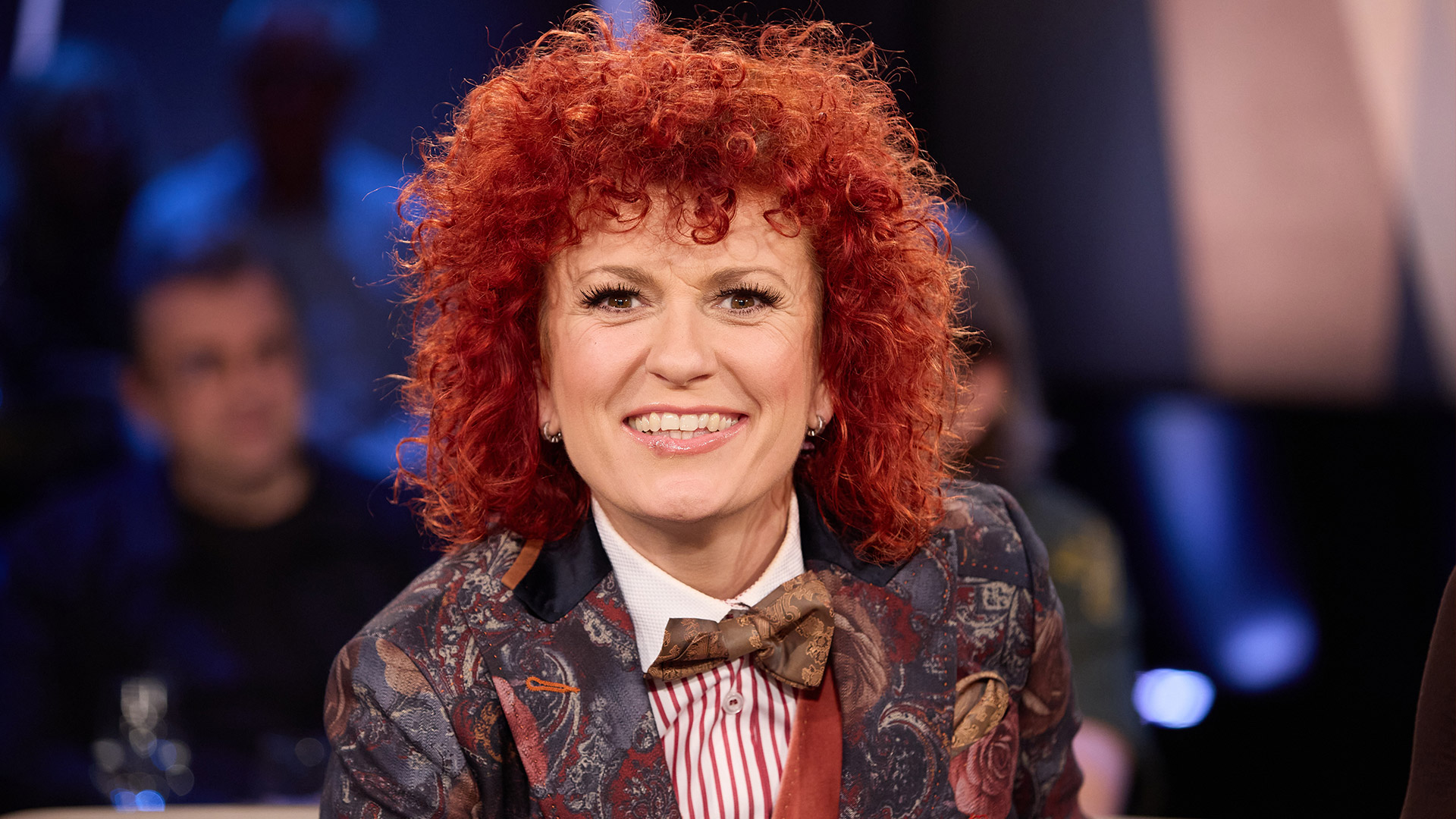 Auf dem Bild ist die Dschungelcamp-Gewinnerin Lucy Diakovska zu sehen. Sie sitzt in einem Studio und lächelt direkt in die Kamera. Lucy trägt ein auffälliges, florales Sakko mit Fliege und gestreiftem Hemd. Ihre roten, lockigen Haare und ihre fröhliche Ausstrahlung stechen hervor. Die Studiobeleuchtung sorgt für eine warme, einladende Atmosphäre.