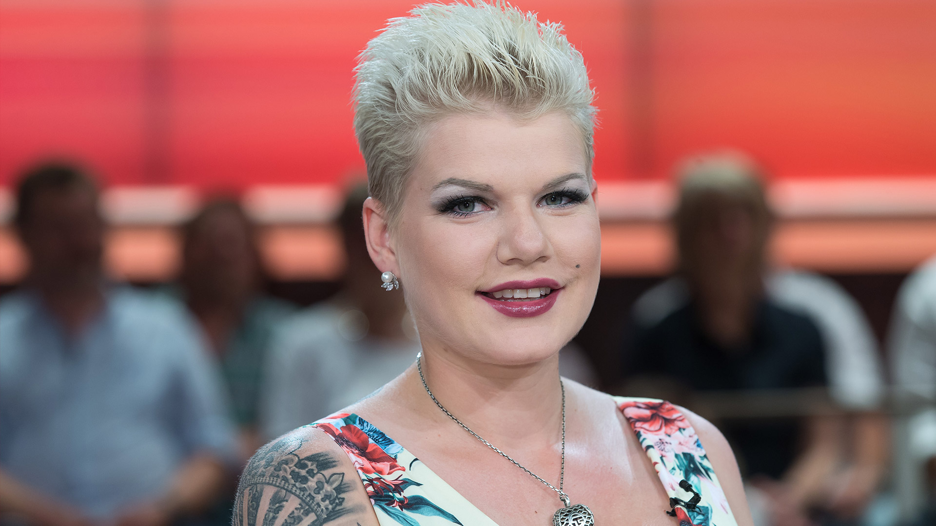 Auf dem Bild ist die Dschungelcamp-Gewinnerin Melanie Müller zu sehen. Sie trägt ein sommerliches, geblümtes Kleid und eine auffällige silberne Herzkette. Ihr blondes, kurzes Haar ist nach hinten gestylt, und ein großes Tattoo ziert ihren rechten Oberarm. Im Hintergrund sitzt ein unscharfes Publikum, die Stimmung wirkt freundlich und entspannt.