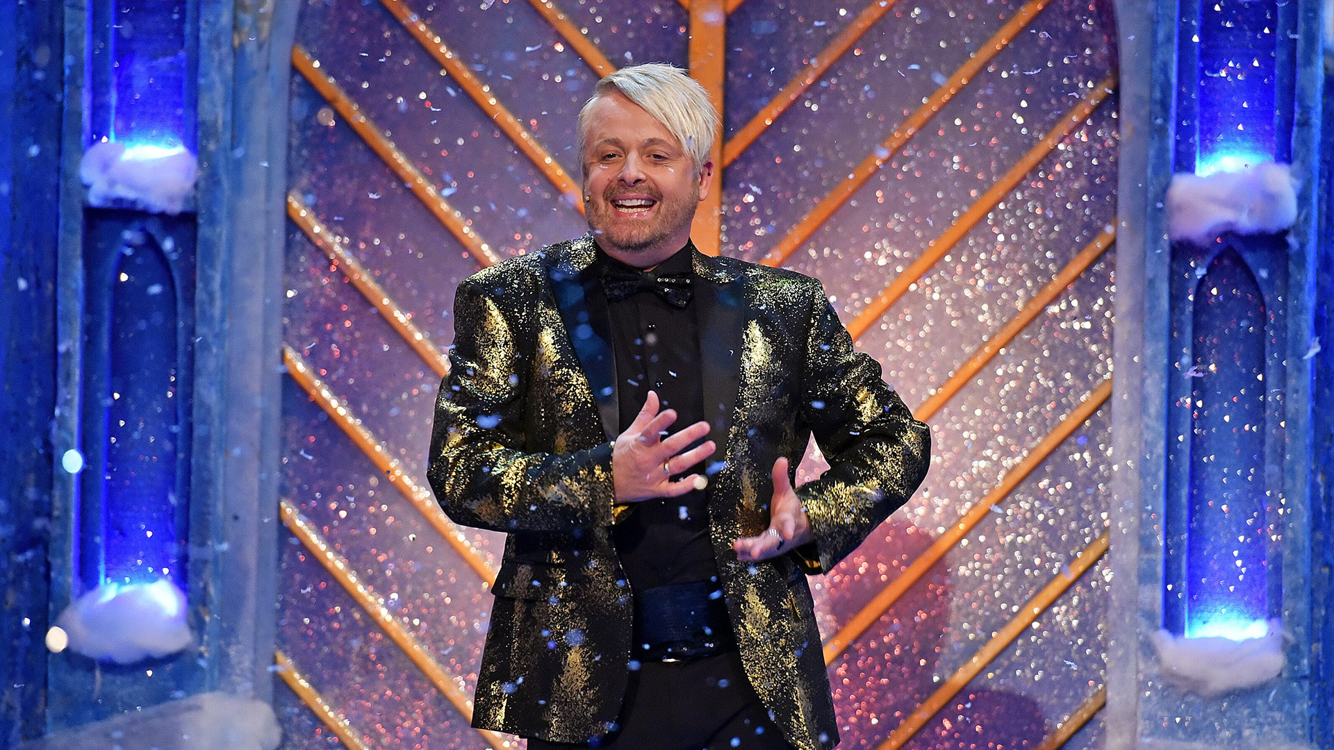 Der Dschungelcamp-Gewinner Ross Antony steht auf einer festlich geschmückten Showbühne mit glitzerndem Hintergrund und Kunstschnee. Er trägt ein auffälliges, golden glänzendes Sakko mit schwarzer Fliege und strahlt mit offenen Gesten in die Kamera. Die winterliche Kulisse unterstreicht die fröhliche und unterhaltsame Atmosphäre des TV-Moments.