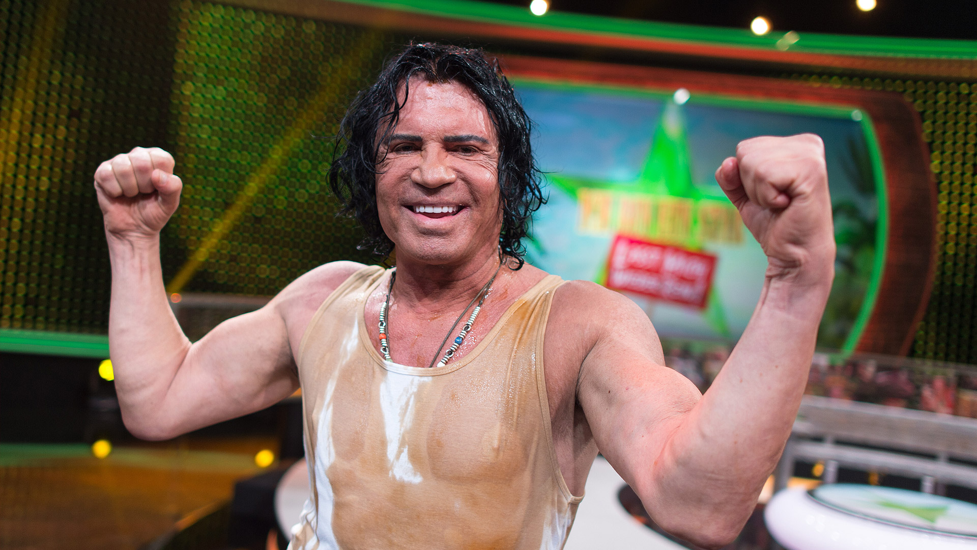 Auf dem Bild ist der Dschungelcamp-Gewinner Costa Cordalis zu sehen. Er steht auf einer Studiobühne, trägt ein verschwitztes, eng anliegendes Tanktop und zeigt beide Arme muskulös und triumphierend in Siegerpose. Sein Gesicht wirkt stolz und zufrieden, im Hintergrund ist das Logo der Dschungelshow zu erkennen. Die Stimmung ist feierlich und energiegeladen.