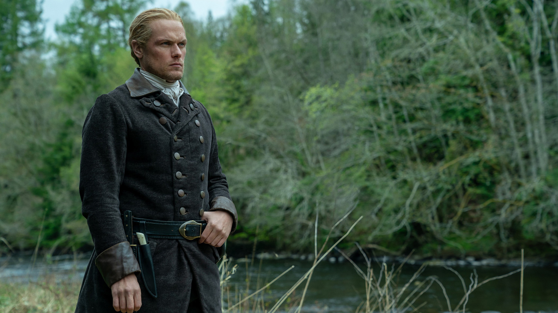 Im Bild zur Outlander-Besetzung steht Sam Heughan als Jamie Fraser in historischer Kleidung mit ernster Miene. Er trägt einen dunklen Mantel mit Knöpfen und einen Gürtel. Hinter ihm erstreckt sich ein dichter, grüner Wald.