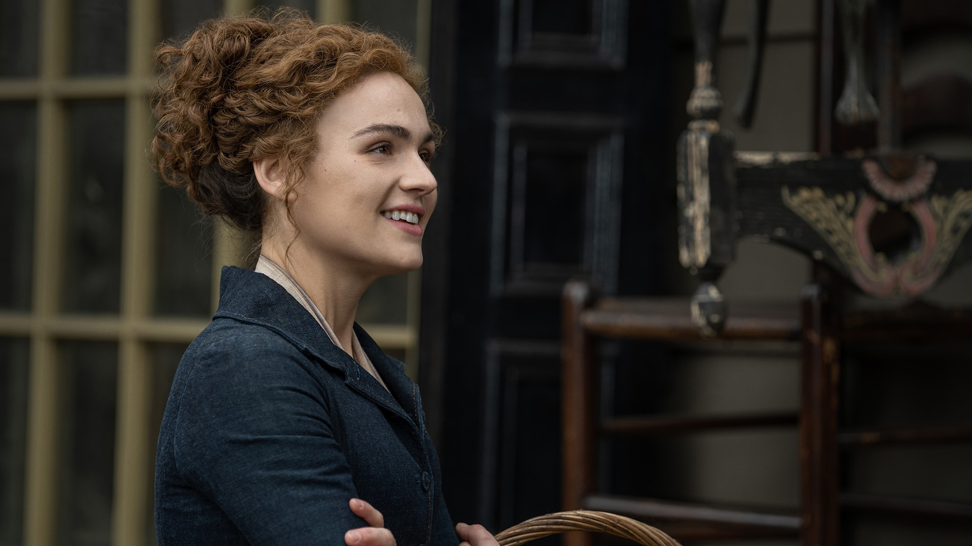 Im Bild zur Outlander-Besetzung ist Sophie Skelton als Brianna Fraser zu sehen. Sie trägt ein schlichtes, dunkles Kleid und lächelt freundlich, während sie seitlich im Profil steht. Ihre rötlich gelockten Haare sind hochgesteckt, und im Hintergrund ist ein historisches Gebäude zu erkennen.
