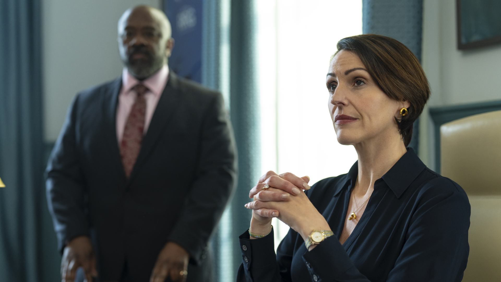 Suranne Jones in Hostage: Eine Frau sitzt mit verschränkten Händen und fokussiertem Blick an einem Schreibtisch, während im Hintergrund ein Mann im Anzug steht.