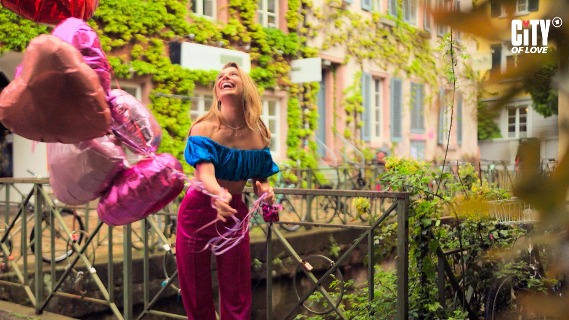 Auf dem Bild in dem Artikel zur ARD-Dating-Show City of Love zieht Lola Weippert mit fröhlichem Lachen mehrere herzförmige Luftballons durch die Luft. Sie steht auf einer kleinen Brücke vor einem Haus mit bewachsenen Fassaden und trägt ein blaues Oberteil sowie eine pinke Hose.