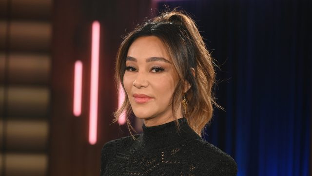 Dieses Symbolbild zu Villa der Versuchung 2026 zeigt Moderatorin Verona Pooth in einem schwarzen, hochgeschlossenen Oberteil mit feinem Muster. Sie steht vor einem Studiolicht-Hintergrund mit dunklen Blau- und Rosatönen. Ihr Blick ist ruhig und selbstbewusst, das Bild vermittelt eine elegante und professionelle Stimmung.