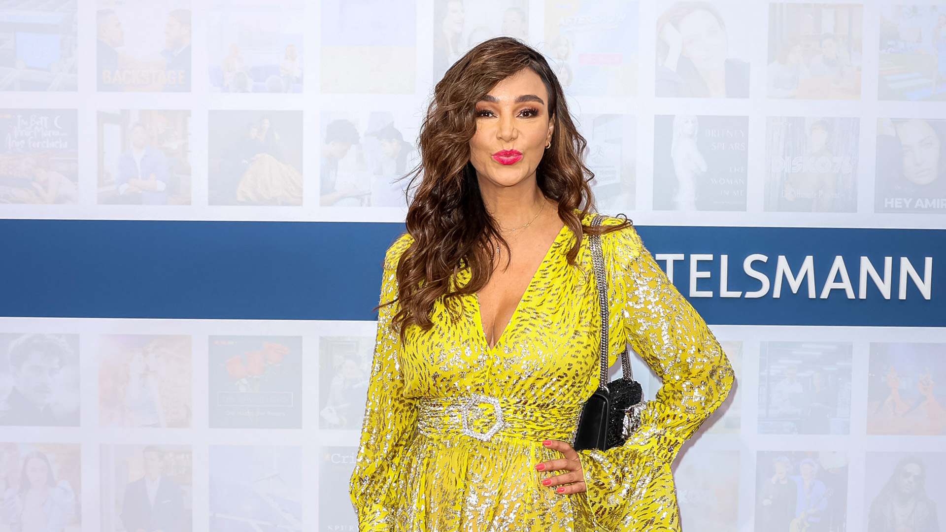 Dieses Symbolbild zu Villa der Versuchung 2026 zeigt Moderatorin Verona Pooth auf einem Event vor einer blauen Bertelsmann-Wand. Sie trägt ein auffälliges, gelbes Kleid mit silbernem Muster und breitem Gürtel. Mit welligem Haar, pinkem Lippenstift und selbstbewusstem Blick strahlt sie Glamour und Präsenz aus – passend zum Reality-TV-Format.