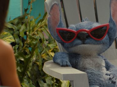 Stitch sitzt auf einem Holzstuhl und trägt eine Sonnenbrille.