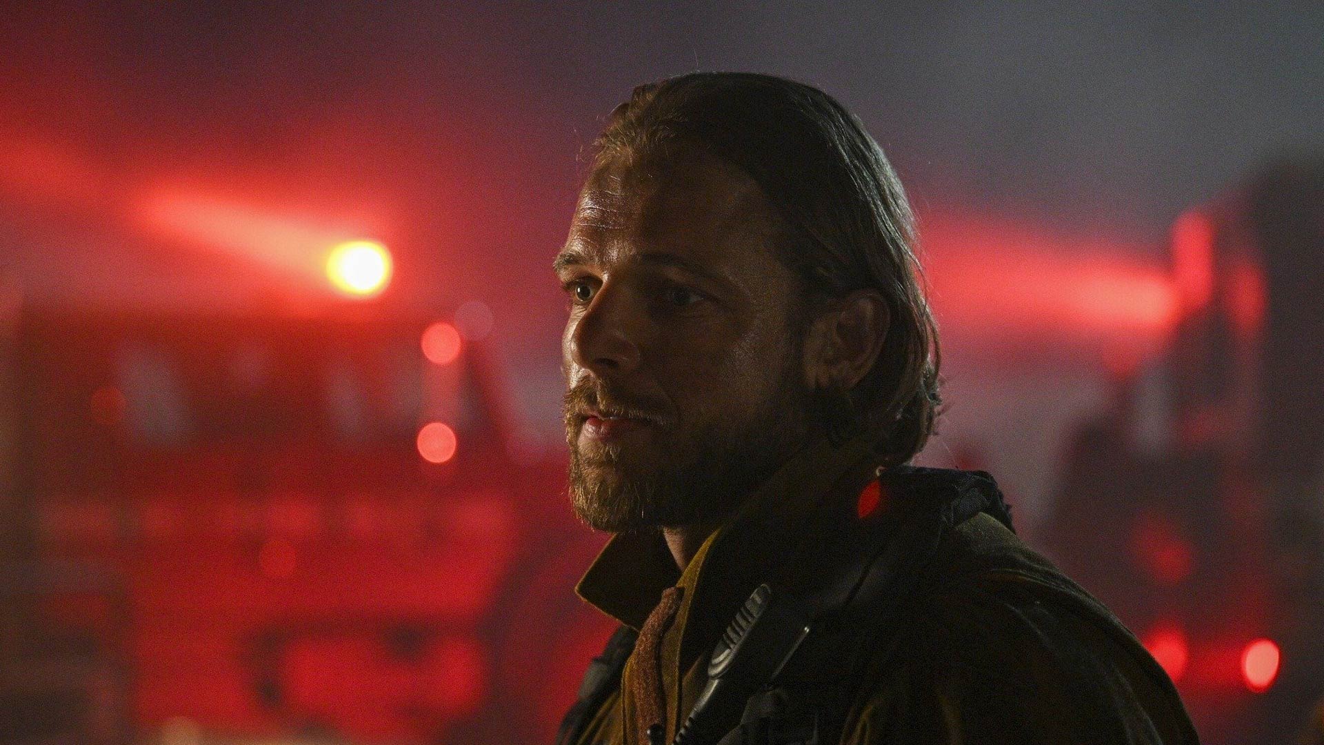 Das Bild in den GigaTV-Highlights im September 2025 ist eine Szene aus Fire Country Staffel 3 und zeigt Bode Donovan, gespielt von Max Thieriot. In feuriger Kulisse mit rot leuchtenden Einsatzfahrzeugen im Hintergrund steht er leicht verschwitzt und ernst im Profil.