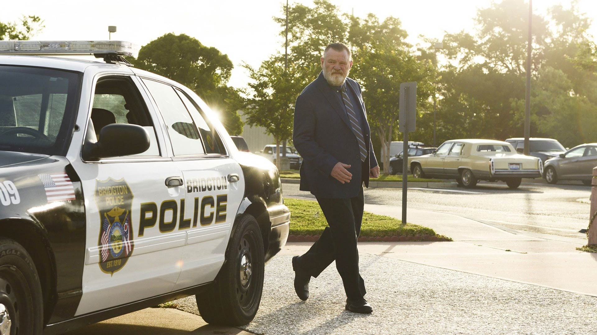 Das Bild in den GigaTV-Highlights im September 2025 zeigt eine Szene aus der Serie Mr. Mercedes. Im Vordergrund steht Detective Bill Hodges, gespielt von Brendan Gleeson, vor einem Polizeiwagen der Stadt Bridgton.