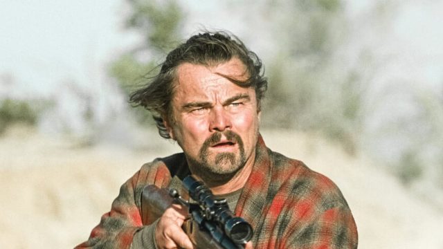 Ein zerzauster und unrasierter Mann steht mit einem Gewehr im Anschlag in einer Wüste.