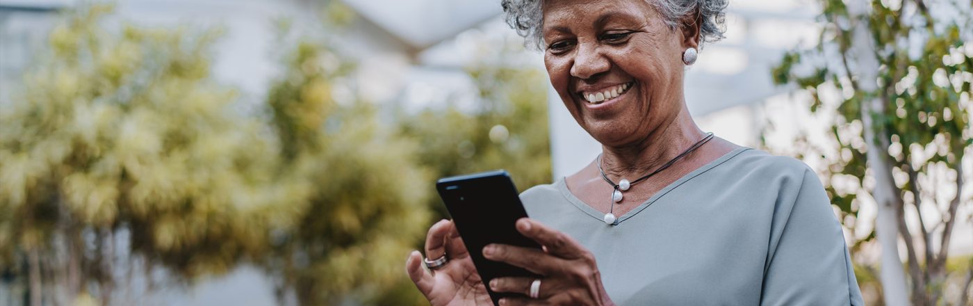 Auf dem Bild in dem Artikel zu den besten Handytarifen für Senior:innen ist eine ältere Frau mit grauen Locken im Freien zu sehen, wie sie entspannt auf ihr Smartphone schaut und lächelt. Sie trägt eine graue Bluse, eine Halskette und ein Smartwatch-Armband.