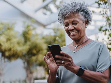 Auf dem Bild in dem Artikel zu den besten Handytarifen für Senior:innen ist eine ältere Frau mit grauen Locken im Freien zu sehen, wie sie entspannt auf ihr Smartphone schaut und lächelt. Sie trägt eine graue Bluse, eine Halskette und ein Smartwatch-Armband.