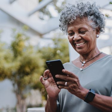 Auf dem Bild in dem Artikel zu den besten Handytarifen für Senior:innen ist eine ältere Frau mit grauen Locken im Freien zu sehen, wie sie entspannt auf ihr Smartphone schaut und lächelt. Sie trägt eine graue Bluse, eine Halskette und ein Smartwatch-Armband.
