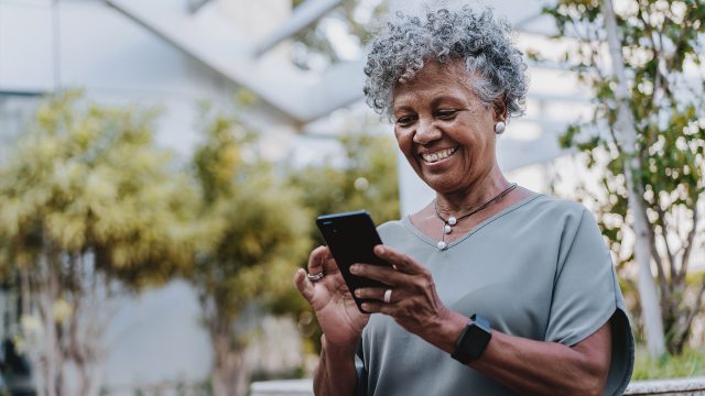 Auf dem Bild in dem Artikel zu den besten Handytarifen für Senior:innen ist eine ältere Frau mit grauen Locken im Freien zu sehen, wie sie entspannt auf ihr Smartphone schaut und lächelt. Sie trägt eine graue Bluse, eine Halskette und ein Smartwatch-Armband.