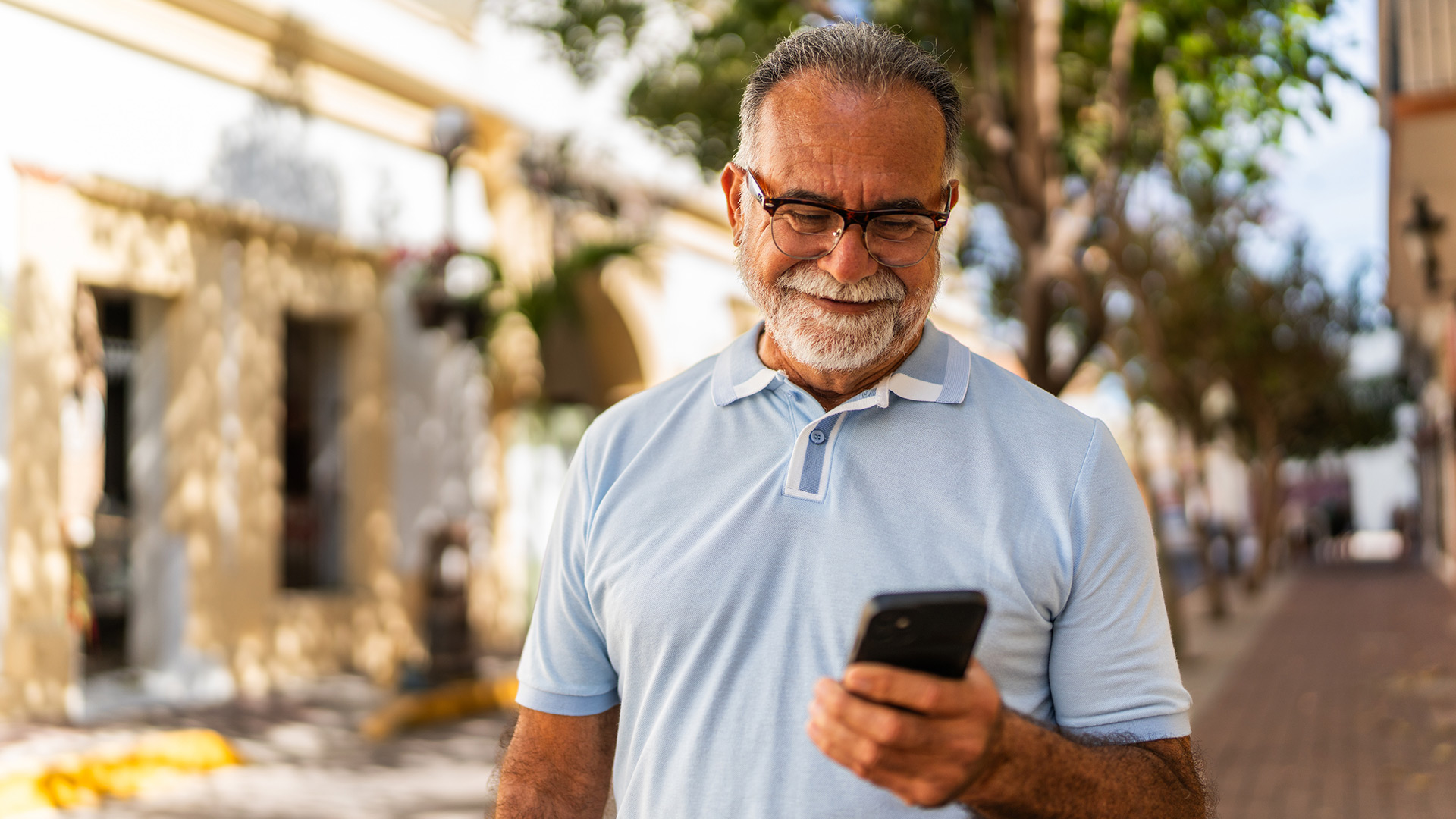 Auf dem Bild in dem Artikel zu den besten Handytarifen für Senior:innen steht ein älterer Mann mit Brille und grauem Bart im Sonnenlicht auf einer Straße. Er trägt ein hellblaues Polohemd und blickt zufrieden auf sein Smartphone.