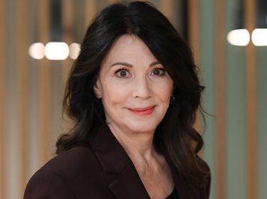 Auf diesem Bild zum Ein fast perfekter Antrag-Film ist Iris Berben zu sehen, die in dem Film eine der Hauptrollen spielt. Sie trägt einen dunkelroten Blazer, hat dunkles, welliges Haar und blickt mit einem dezenten Lächeln in die Kamera. Der Hintergrund ist unscharf mit vertikalen Holzstreben und warmem Licht.