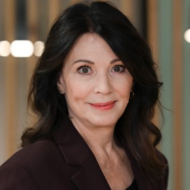 Auf diesem Bild zum Ein fast perfekter Antrag-Film ist Iris Berben zu sehen, die in dem Film eine der Hauptrollen spielt. Sie trägt einen dunkelroten Blazer, hat dunkles, welliges Haar und blickt mit einem dezenten Lächeln in die Kamera. Der Hintergrund ist unscharf mit vertikalen Holzstreben und warmem Licht.