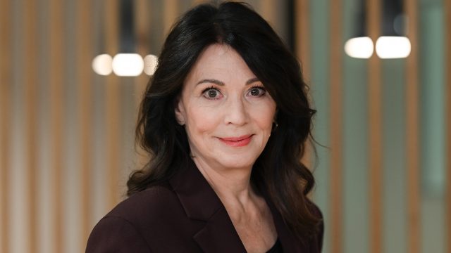 Auf diesem Bild zum Ein fast perfekter Antrag-Film ist Iris Berben zu sehen, die in dem Film eine der Hauptrollen spielt. Sie trägt einen dunkelroten Blazer, hat dunkles, welliges Haar und blickt mit einem dezenten Lächeln in die Kamera. Der Hintergrund ist unscharf mit vertikalen Holzstreben und warmem Licht.