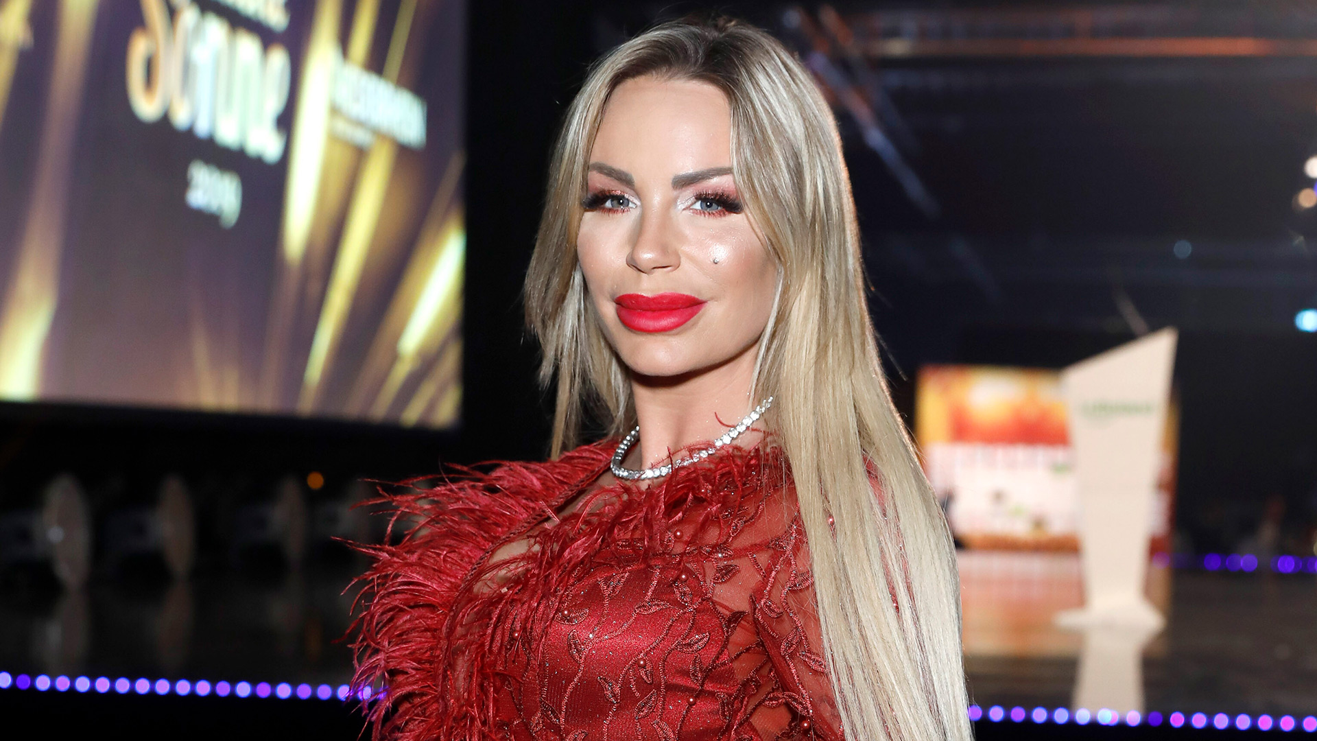 Auf dem Bild in dem Artikel zu Promis unter Palmen 2026 ist Gina-Lisa Lohfink in einem roten Kleid mit Federn und Glitzerelementen zu sehen. Sie trägt Make-up und roten Lippenstift, ihr blondes Haar fällt offen über die Schultern.