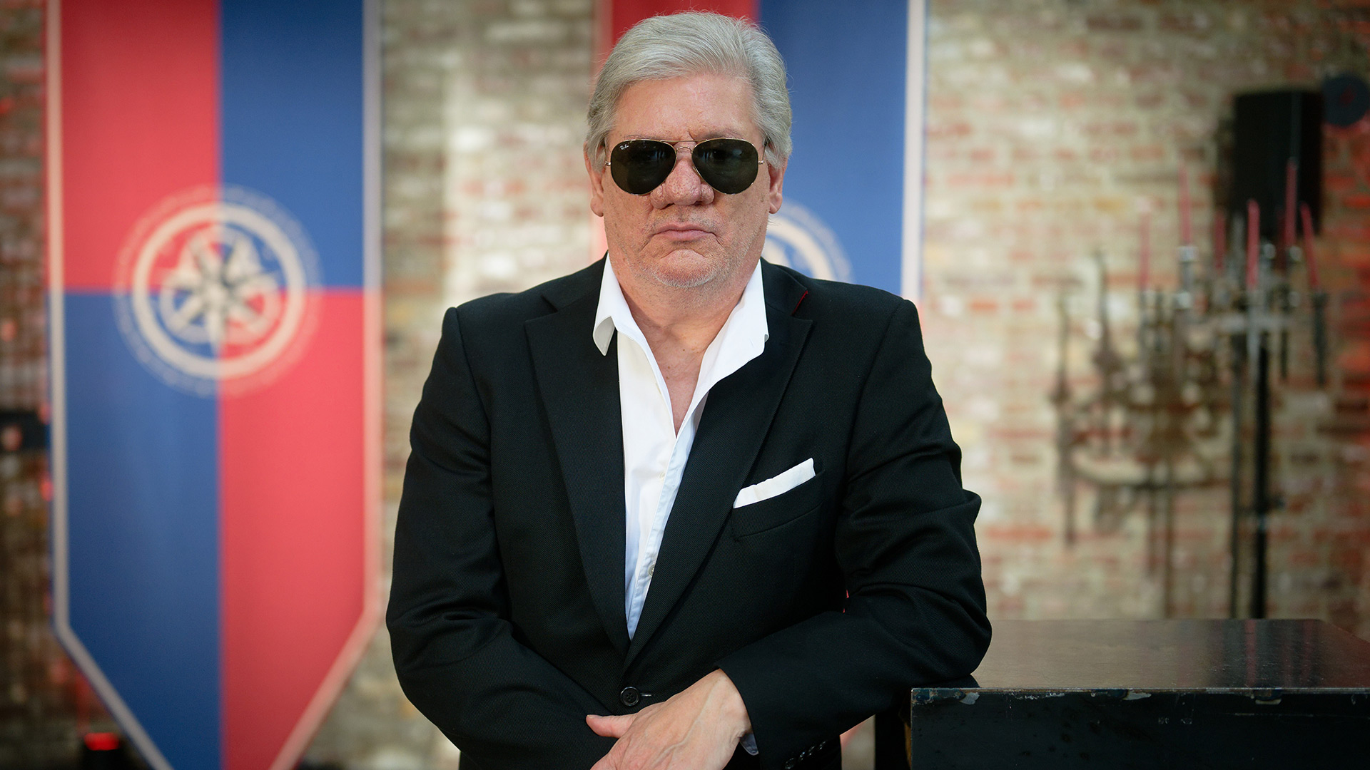 Auf dem Bild in dem Artikel zu Promis unter Palmen 2026 ist Claude-Oliver Rudolph in einem schwarzen Anzug mit weißem Hemd und Sonnenbrille zu sehen. Er steht mit verschränkten Armen vor einer rustikalen Backsteinwand.