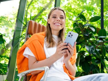 Auf dem Bild in dem Artikel zur Samsung Garantie sitzt eine junge Frau entspannt in einem modernen, hellen Raum mit vielen Pflanzen und hält ein Smartphone in der Hand.