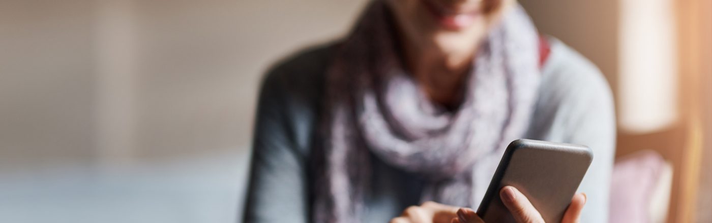 Auf dem Bild zum Thema Smartphones für Senior:innen ist eine ältere Frau mit grauen Haaren und Brille zu sehen, die entspannt auf einem Stuhl sitzt und lächelnd ein Smartphone bedient. Sie trägt einen Schal und wirkt zufrieden und konzentriert. Die Stimmung ist ruhig und positiv, das Licht ist warm und freundlich.