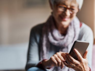 Auf dem Bild zum Thema Smartphones für Senior:innen ist eine ältere Frau mit grauen Haaren und Brille zu sehen, die entspannt auf einem Stuhl sitzt und lächelnd ein Smartphone bedient. Sie trägt einen Schal und wirkt zufrieden und konzentriert. Die Stimmung ist ruhig und positiv, das Licht ist warm und freundlich.