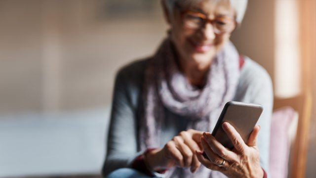 Auf dem Bild zum Thema Smartphones für Senior:innen ist eine ältere Frau mit grauen Haaren und Brille zu sehen, die entspannt auf einem Stuhl sitzt und lächelnd ein Smartphone bedient. Sie trägt einen Schal und wirkt zufrieden und konzentriert. Die Stimmung ist ruhig und positiv, das Licht ist warm und freundlich.