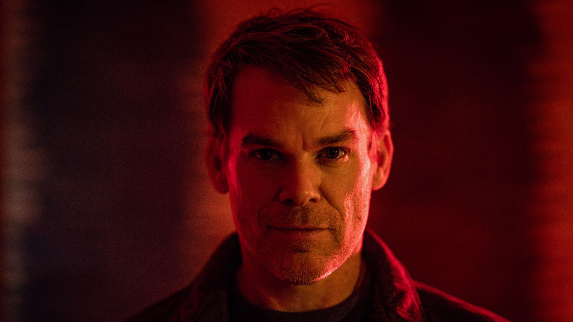 Auf dem Bild zur Serie Dexter Wiedererwachen ist Dexter Morgan zu sehen, gespielt von Michael C. Hall. Er steht im Mittelpunkt des Bildes, sein Gesicht wird von dramatischem, rötlichem Licht betont, was eine düstere und intensive Stimmung erzeugt. Die Szene wirkt geheimnisvoll und emotional aufgeladen
