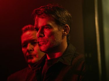 Auf dem Bild zur Serie Dexter Wiedererwachen ist vorne Dexter Morgan, gespielt von Michael C. Hall, mit ernstem Blick zu sehen. Hinter ihm steht Harry Morgan, gespielt von James Remar, im Schatten. Beide wirken angespannt, das rote Licht verstärkt die bedrohliche Atmosphäre.