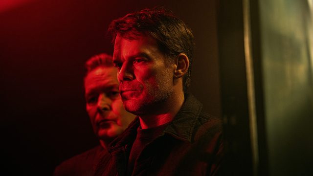Auf dem Bild zur Serie Dexter Wiedererwachen ist vorne Dexter Morgan, gespielt von Michael C. Hall, mit ernstem Blick zu sehen. Hinter ihm steht Harry Morgan, gespielt von James Remar, im Schatten. Beide wirken angespannt, das rote Licht verstärkt die bedrohliche Atmosphäre.