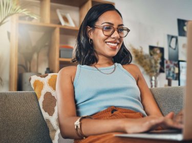 Eine Frau sitzt mit einem Laptop auf dem Schoß auf einer Couch.