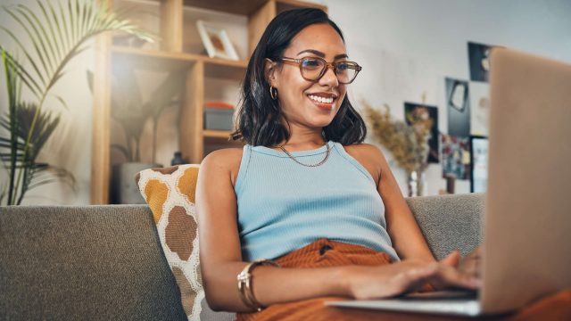 Eine Frau sitzt mit einem Laptop auf dem Schoß auf einer Couch.