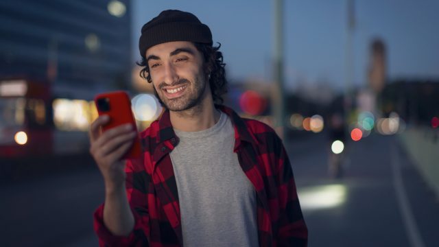 Im Bild zur Garantie bei Apple siehst du einen jungen Mann mit dunklen Locken und roter Handyhülle, der in der Abenddämmerung lächelnd auf sein iPhone schaut.