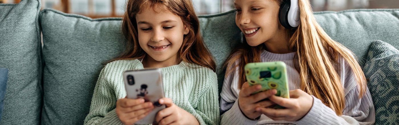 Auf dem Bild zum Thema Smartphones für Kinder sitzen zwei Mädchen fröhlich nebeneinander auf einem Sofa. Beide halten jeweils ein Smartphone in der Hand, eines mit bunter Hülle, das andere mit Tiermotiv. Das Mädchen rechts trägt zusätzlich große weiße Kopfhörer und lächelt ihre Freundin an. Die Szene wirkt entspannt und spielerisch.