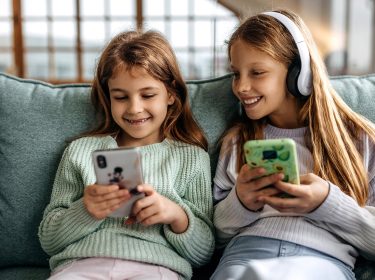 Auf dem Bild zum Thema Smartphones für Kinder sitzen zwei Mädchen fröhlich nebeneinander auf einem Sofa. Beide halten jeweils ein Smartphone in der Hand, eines mit bunter Hülle, das andere mit Tiermotiv. Das Mädchen rechts trägt zusätzlich große weiße Kopfhörer und lächelt ihre Freundin an. Die Szene wirkt entspannt und spielerisch.