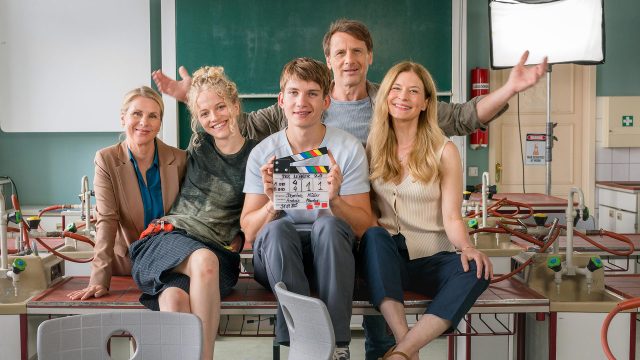 Auf dem Bild in dem Artikel zur Der Lehrer-Besetzung sind fünf Personen, die lächelnd auf einem Tisch im Klassenzimmer sitzen zu sehen. In der Mitte hält eine junge Person eine Filmklappe mit dem Titel „Der Lehrer 2.0“.