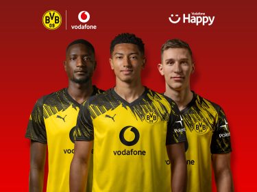 Auf dem Bild in dem Artikel zum BVB-Gewinnspiel stehen drei Spieler von Borussia Dortmund nebeneinander vor einem roten Hintergrund. Sie tragen das gelb-schwarze Trikot mit dem Vodafone-Logo auf der Brust. Über ihnen sind die Logos von BVB, Vodafone und Vodafone Happy zu sehen.