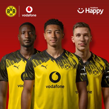 Auf dem Bild in dem Artikel zum BVB-Gewinnspiel stehen drei Spieler von Borussia Dortmund nebeneinander vor einem roten Hintergrund. Sie tragen das gelb-schwarze Trikot mit dem Vodafone-Logo auf der Brust. Über ihnen sind die Logos von BVB, Vodafone und Vodafone Happy zu sehen.