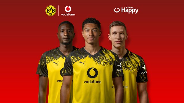Auf dem Bild in dem Artikel zum BVB-Gewinnspiel stehen drei Spieler von Borussia Dortmund nebeneinander vor einem roten Hintergrund. Sie tragen das gelb-schwarze Trikot mit dem Vodafone-Logo auf der Brust. Über ihnen sind die Logos von BVB, Vodafone und Vodafone Happy zu sehen.