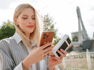 Frau steht in Großstadt in der Nähe einer Brücke und hält in einer Hand ein iPhone, in der anderen eine Powerbank