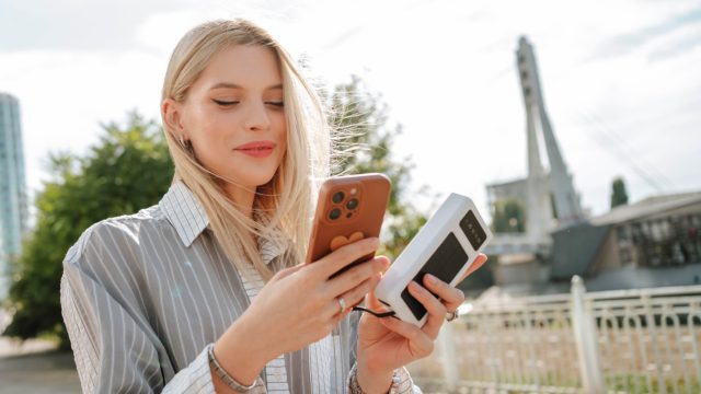 Frau steht in Großstadt in der Nähe einer Brücke und hält in einer Hand ein iPhone, in der anderen eine Powerbank