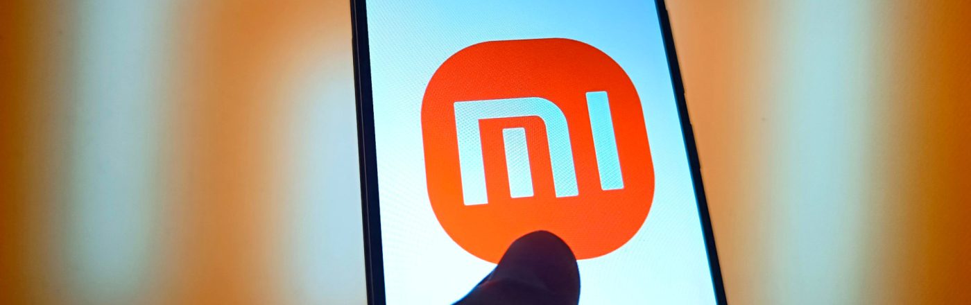 Smartphone mit Mi-Logo und einem Daumen auf dem Display vor orangefarbenem Hintergrund