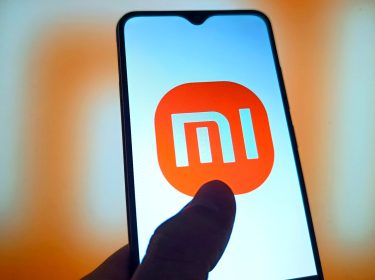 Smartphone mit Mi-Logo und einem Daumen auf dem Display vor orangefarbenem Hintergrund