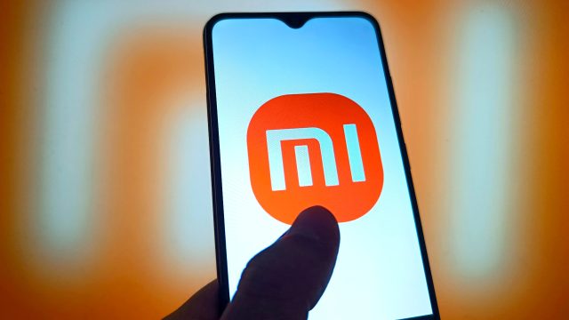 Smartphone mit Mi-Logo und einem Daumen auf dem Display vor orangefarbenem Hintergrund